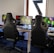A row of gaming stations equipped with large monitors, gaming chairs, keyboards, and headphones. The setup is organized and ready for gaming, featuring a colorful screen display. The room has a modern and tech-savvy ambiance.