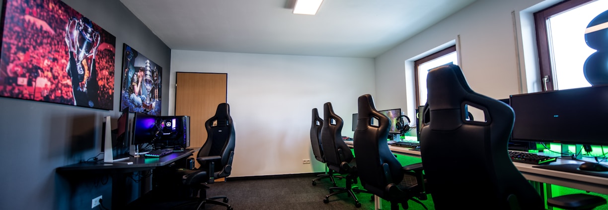 A well-lit gaming room features multiple gaming stations with high-backed chairs and monitors. The room has a minimalist setup with green and black color tones. Large posters adorn the walls, and the room appears clean and organized.