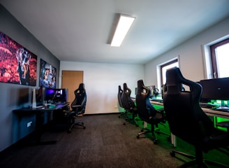 A well-lit gaming room features multiple gaming stations with high-backed chairs and monitors. The room has a minimalist setup with green and black color tones. Large posters adorn the walls, and the room appears clean and organized.