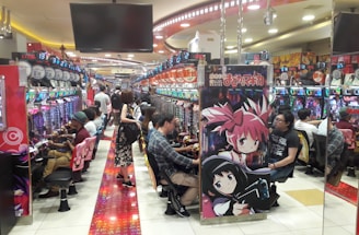 A bustling arcade filled with people playing various gaming machines. Brightly colored displays and animated characters are prominent on posters and screens. The atmosphere is lively with patrons seated in rows, focusing on the games. Overhead signage and electronic scoreboards are visible, contributing to the busy and vibrant environment.