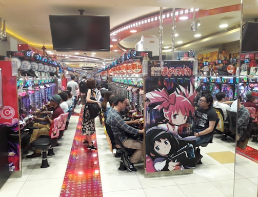 A bustling arcade filled with people playing various gaming machines. Brightly colored displays and animated characters are prominent on posters and screens. The atmosphere is lively with patrons seated in rows, focusing on the games. Overhead signage and electronic scoreboards are visible, contributing to the busy and vibrant environment.