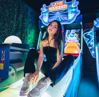 A person sitting on an arcade game machine platform with neon lights in the background. The arcade game is called 'Lane Master' and features a colorful design with bowling-themed imagery.