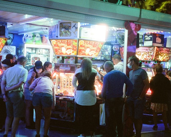 A group of people standing in front of an illuminated amusement arcade, engaging with various gaming machines. The environment is bustling with activity, and colorful lights create a vibrant atmosphere. The ceiling is decorated with large, playful images of dice and other game-related graphics, enhancing the lively mood.