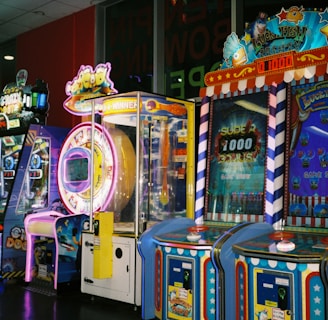 A row of brightly colored arcade game machines is arranged in a line against the wall. Each machine features vibrant lights and designs, with themes ranging from claw games to game show-inspired setups. The area has a playful and inviting atmosphere, suitable for entertainment or amusement centers.