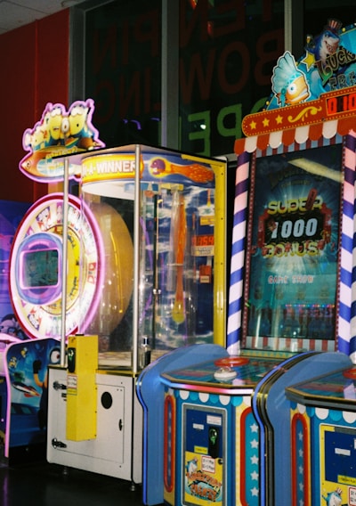 A row of brightly colored arcade game machines is arranged in a line against the wall. Each machine features vibrant lights and designs, with themes ranging from claw games to game show-inspired setups. The area has a playful and inviting atmosphere, suitable for entertainment or amusement centers.