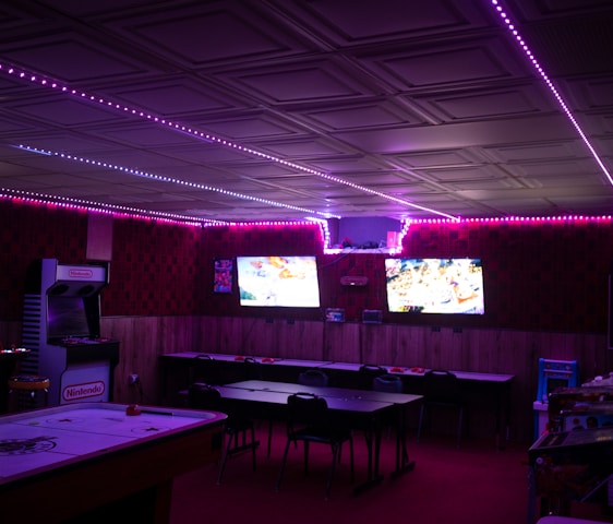 A dimly lit room with vibrant purple LED strip lights on the ceiling, featuring multiple gaming stations. There are two large screens showing video games, along with various arcade machines, including a Nintendo arcade cabinet. An air hockey table is positioned in the foreground.