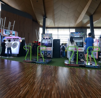A vibrant arcade setting with several gaming machines lined up, including dance and rhythm game setups. People are engaged with the games, absorbed in their activities. The flooring is wooden, and the ceiling has a geometric design. Natural light filters through large windows, creating a lively atmosphere.