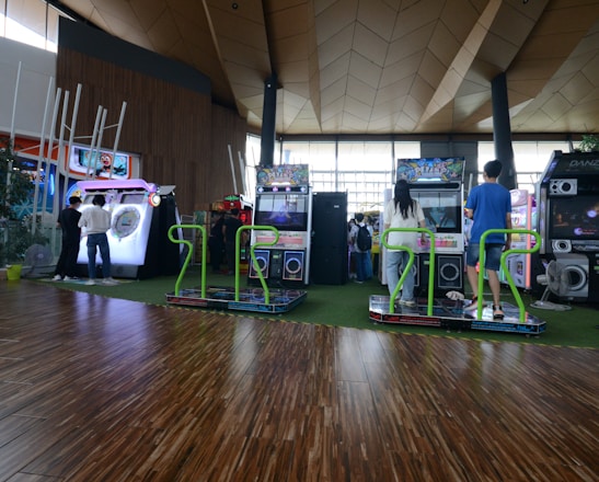 A vibrant arcade setting with several gaming machines lined up, including dance and rhythm game setups. People are engaged with the games, absorbed in their activities. The flooring is wooden, and the ceiling has a geometric design. Natural light filters through large windows, creating a lively atmosphere.