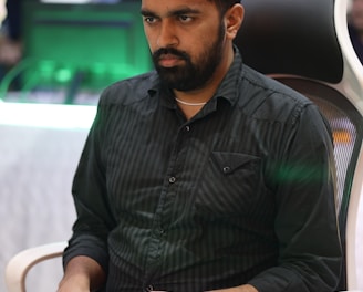 A person is seated in a chair, focused intently on something, with hands positioned on a gaming console or arcade controller. The setup appears to be indoors, with visible screens and lighting in the background, suggesting an electronic gaming environment.