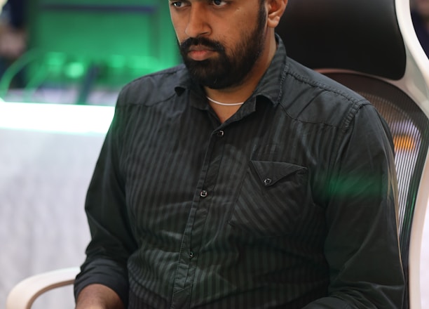 A person is seated in a chair, focused intently on something, with hands positioned on a gaming console or arcade controller. The setup appears to be indoors, with visible screens and lighting in the background, suggesting an electronic gaming environment.